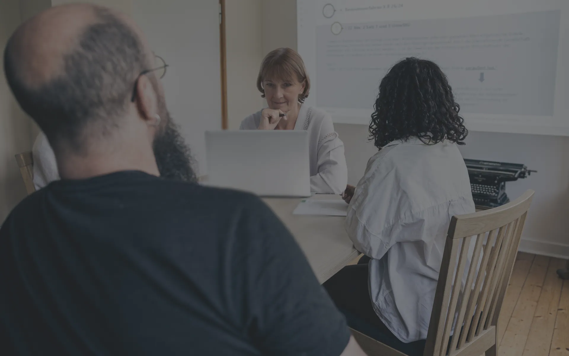 Ein Meeting unter Steuerberatern wird an einem Schreibtisch durchgeführt, während im Hintergrund eine Leinwand zu sehen ist.