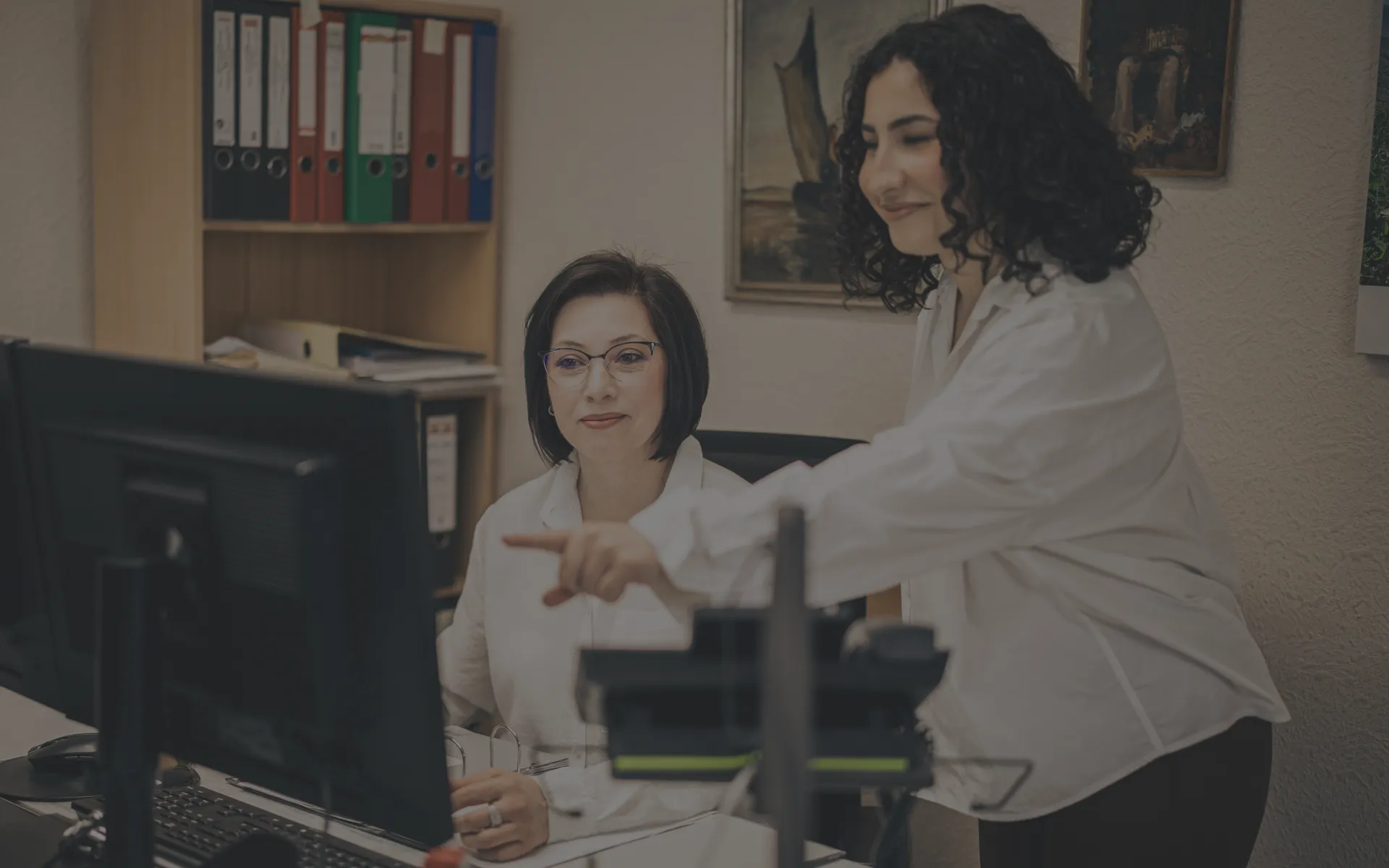 In einem Steuerbüro zeigt eine Kollegin einer anderen freundlich etwas auf einem Monitor.